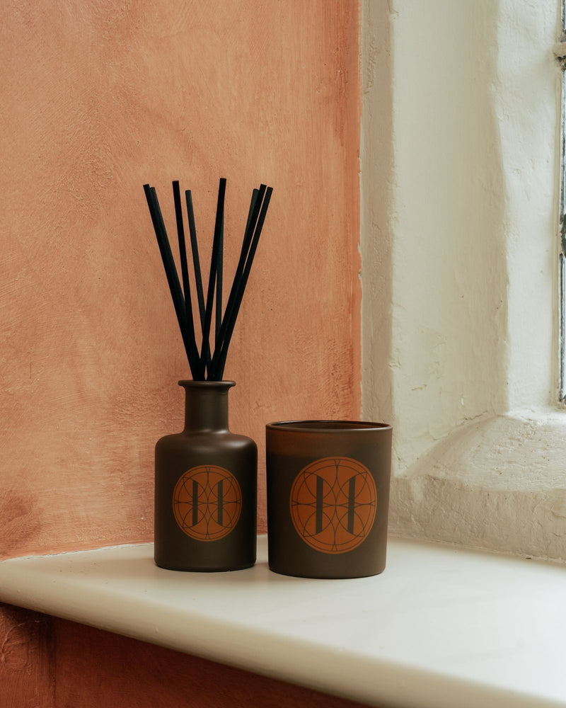 Two dark brown ceramic containers with candle and diffuser with black reeds on a windowsill.