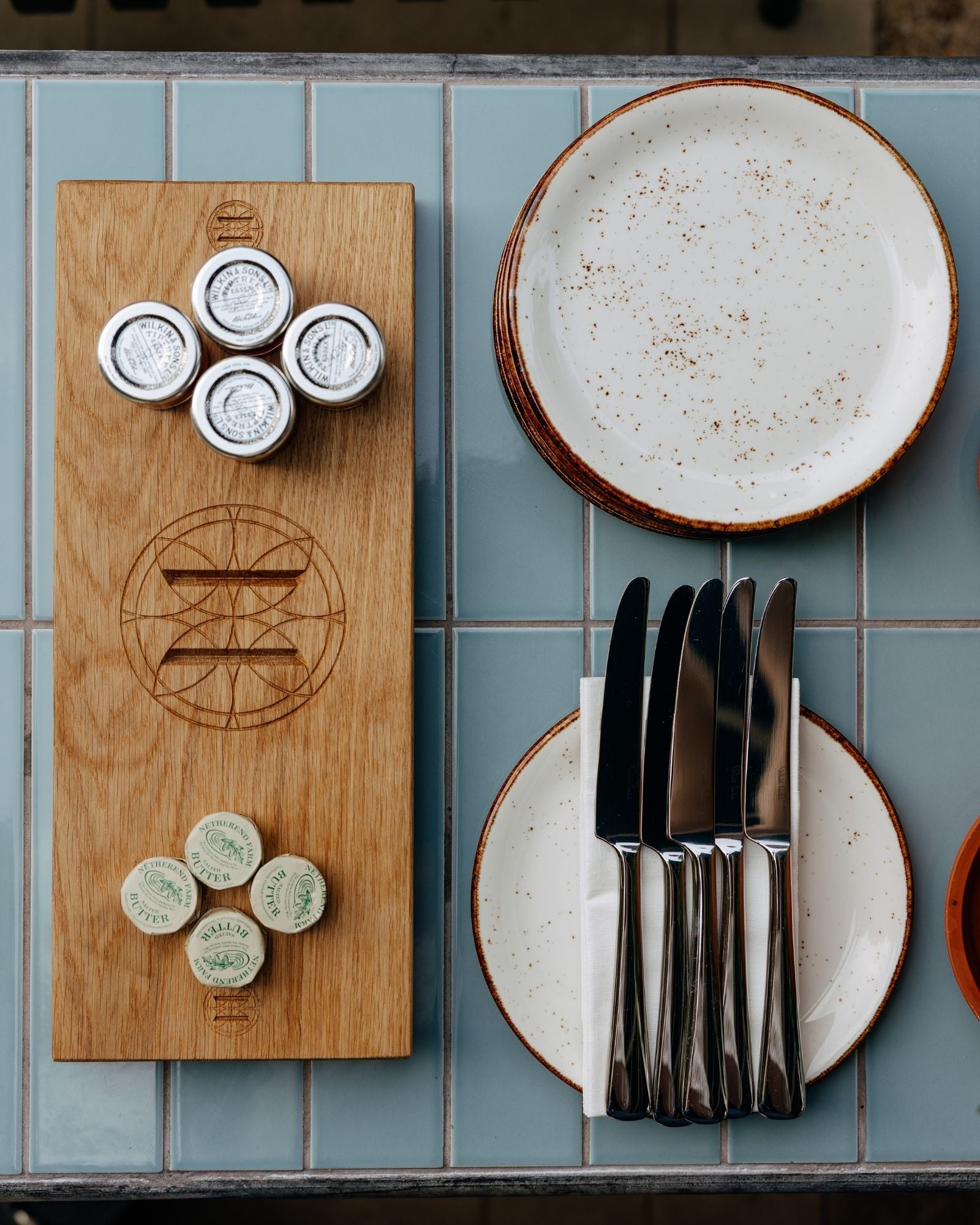 Wooden cutting board with engraved design, ceramic plates, and cutlery on a tiled surface.