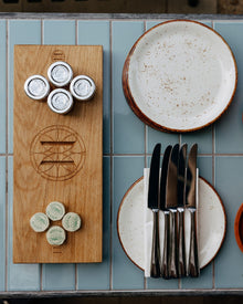 Wooden cutting board with engraved design, ceramic plates, and cutlery on a tiled surface.