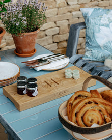 Wooden board with pastries, jam jars, and cutlery on a table with a stone wall background.