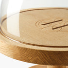 Close-up of oak pedestal cake stand with glass dome and Eleven Bibury engraved logo on a white background.