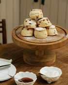 Stack of scones on a wooden stand with jam and cream on a table.
