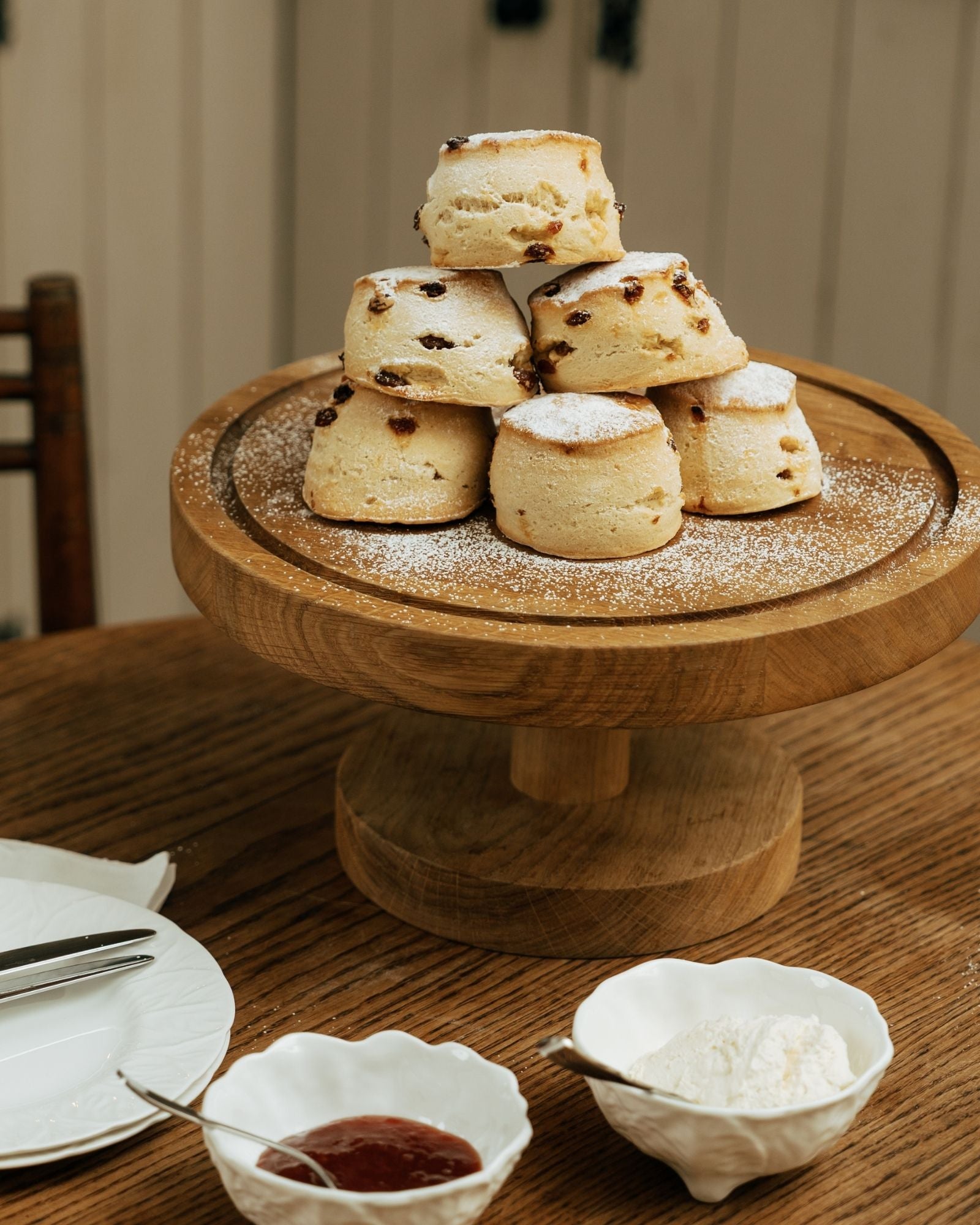 Stack of scones on a wooden stand with jam and cream on a table.