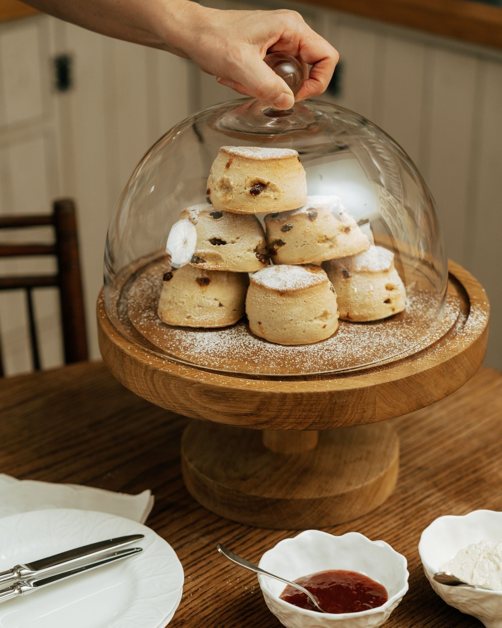 Wooden cake stand with glass dome holding scones on a wooden table.