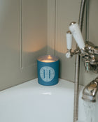 Blue Rack Isle candle with a Eleven Bibury logo on a bath counter next to a shower head.