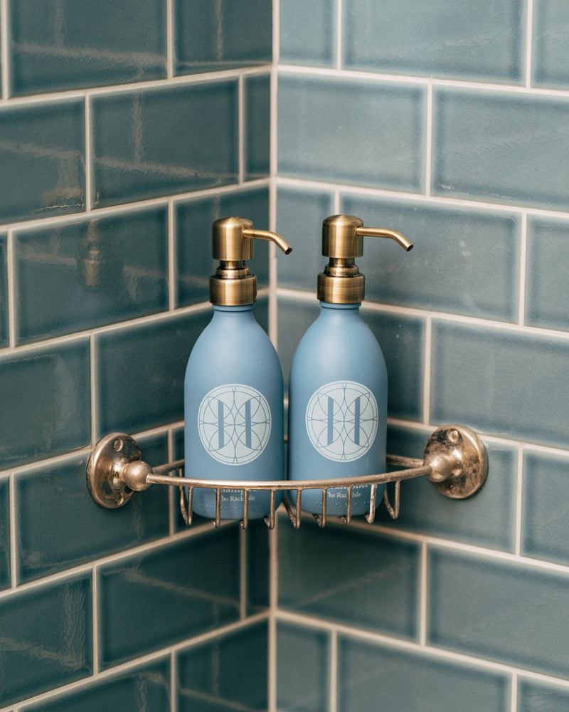Two blue shampoo and conditioner dispensers with gold pumps on a wall-mounted rack against a tiled bathroom wall.