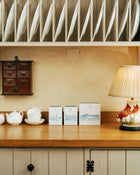 Wooden shelf with tea, coffee and hot chocolate items and a lamp in a cozy kitchen setting.