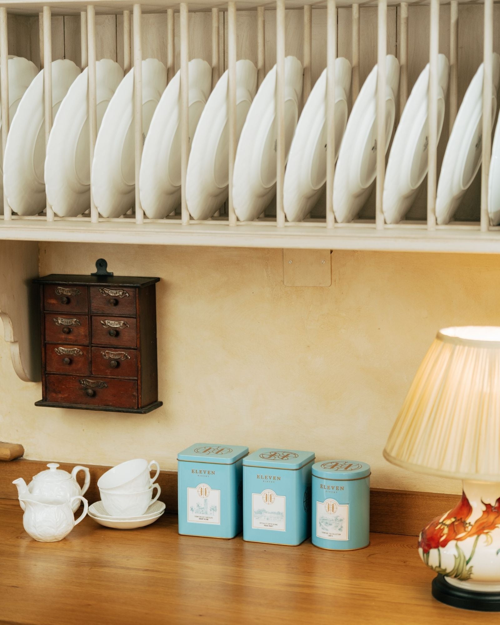 Dish rack with white plates above a wooden shelf with tea, coffee, hot chocolate tins on counter.