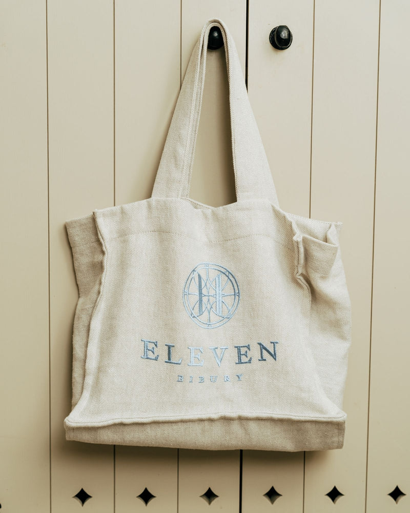 Beige tote bag with 'Eleven Bibury' branding hanging on a wooden cabinet.